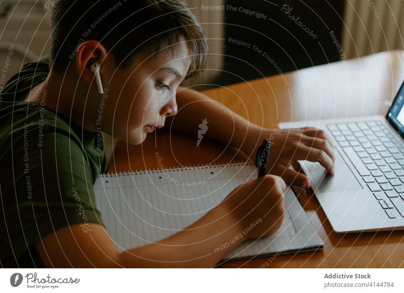 Concentrated kid writing in notebook during online studies at home boy homework assignment laptop write information distance wireless earphones study education