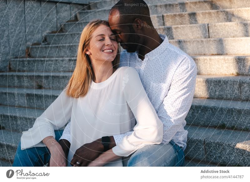 Tender multiracial couple resting on street stairs and hugging bonding in love romantic content tender sensual eyes closed cuddle happy cheerful fondness