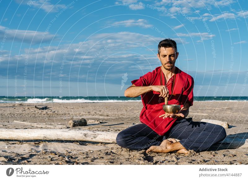 Calm man relaxing while playing singing bowl on beach tibetan meditate half lotus ocean peaceful asana ayurveda male casual young tradition culture buddhism zen