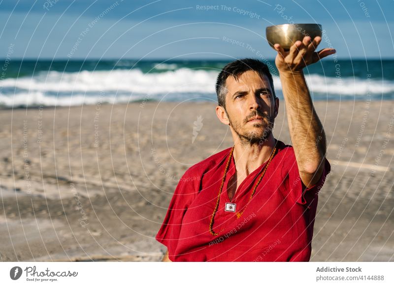Peaceful man using Tibetan singing bowl for stress relief on beach play tibetan ocean meditate tradition culture relax cloudless blue sky male shirt sand sea