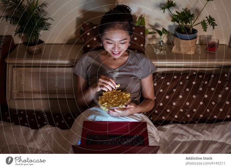 Content Asian lady watching movie on laptop while eating popcorn woman enjoy relax domestic using gadget device internet film comfort cheerful night netbook