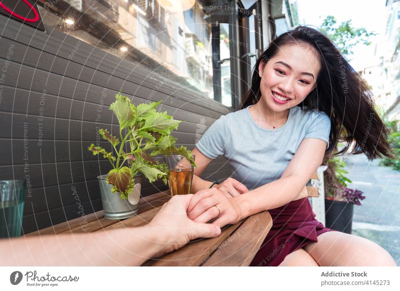 Joyful Asian woman talking with crop unrecognizable friend in cafe communicate beverage glass excited laugh together free time friendship legs crossed