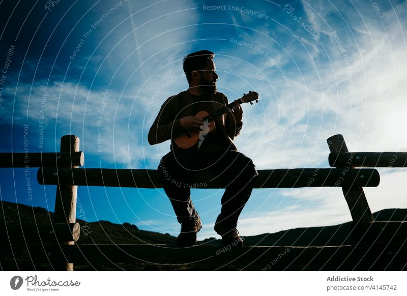 Bearded man playing ukulele in countryside fence sky silhouette cloudy music puerto de la morcuera spain male travel serious vacation trip nature rest journey