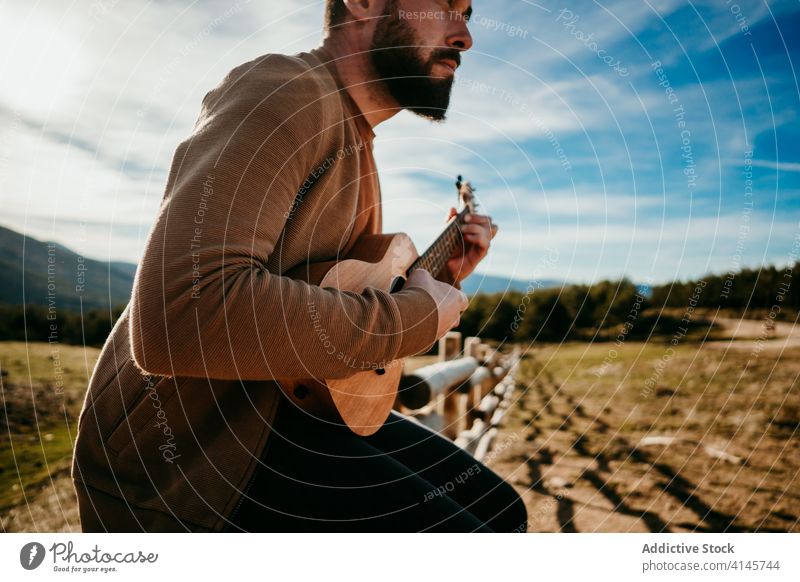 Anonymous bearded man playing ukulele in countryside fence sky cloudy music puerto de la morcuera spain male travel vacation trip nature rest journey sunny