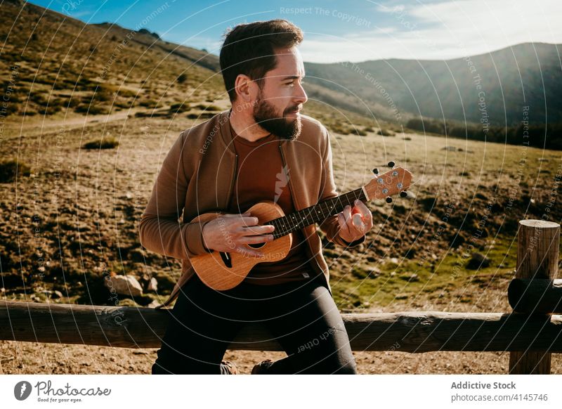 Bearded man playing ukulele in countryside fence sky cloudy music puerto de la morcuera spain male travel serious vacation trip nature rest journey sunny