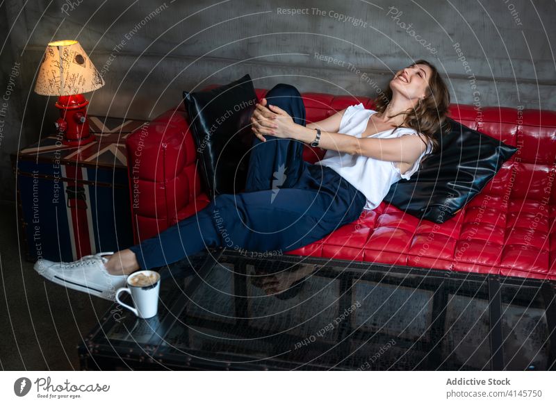 Young woman lying down on leather sofa rest style cheerful comfort cozy relax happy trendy modern female young couch creative design interior lifestyle beverage