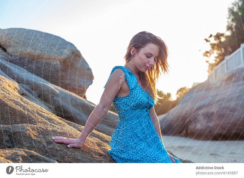 Stylish young woman leaning on huge cliff on summer day relax lean on rock traveler enjoy happy touch hair seaside vacation trip female dress style trendy