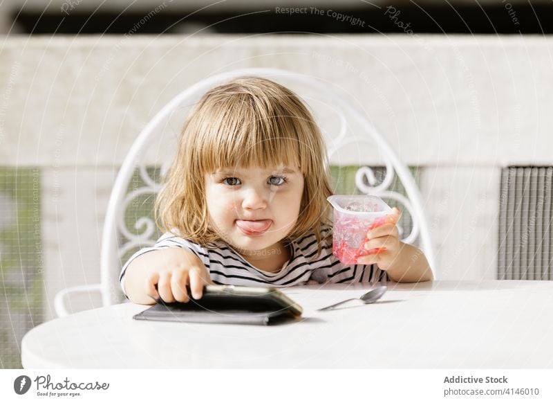 Kid eating jelly and using smartphone on terrace child watch cartoon little girl entertain summer backyard table cute childhood sit kid rest adorable relax