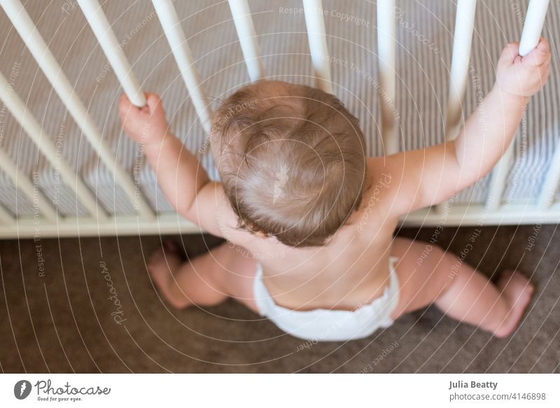 Looking down at a toddler lowering self to sit while holding onto crib rails; baby learning to stand and sit independently infant child childhood 6-12 months