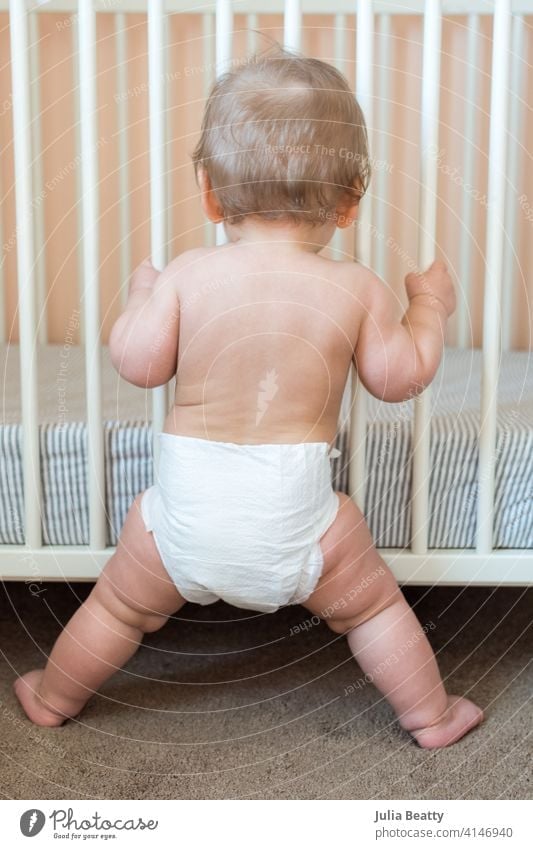 Toddler standing with legs apart while holding onto to crib rails: nursery with peach, gray and white decor baby infant child childhood 6-12 months pull up