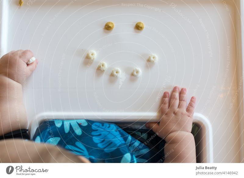 Baby led weaning made fun: smiley face of cereal puffs on a high chair tray with baby's hand resting nearby infant child 6-12 months old finger food yogurt
