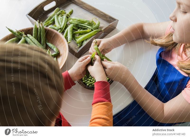 Crop children peeling pile of pea pods at home help fresh vegetarian bowl together sibling raw childhood uncooked product wooden round table green crunchy