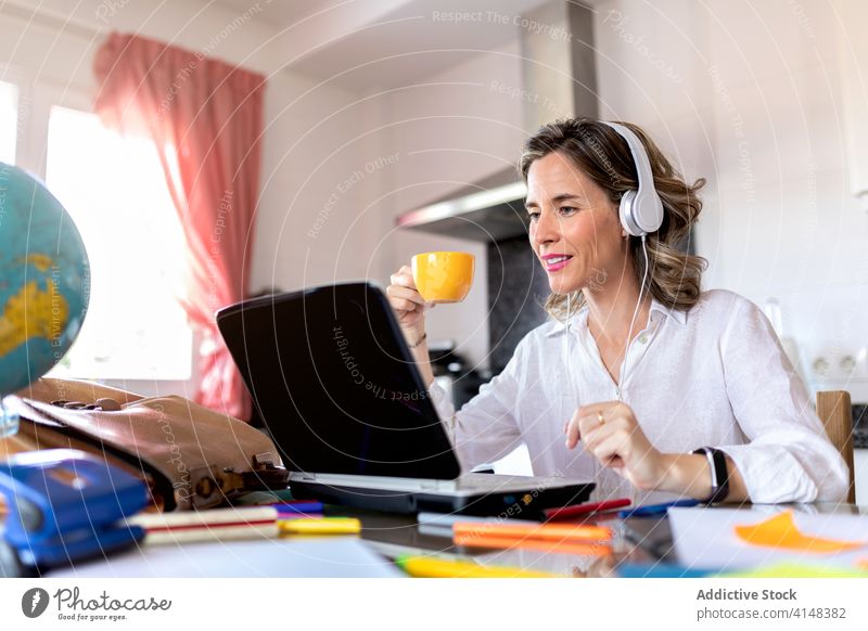 Positive woman in headset having video call on laptop indoors cup hot drink free time speak smile using gadget device netbook conversation video chat table