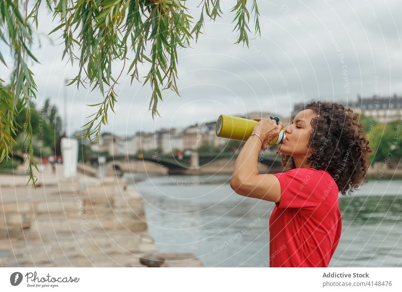 Thirsty ethnic sportswoman drinking water after working out near river thirst bottle headset sportswear break tree using gadget headphones device urban plastic