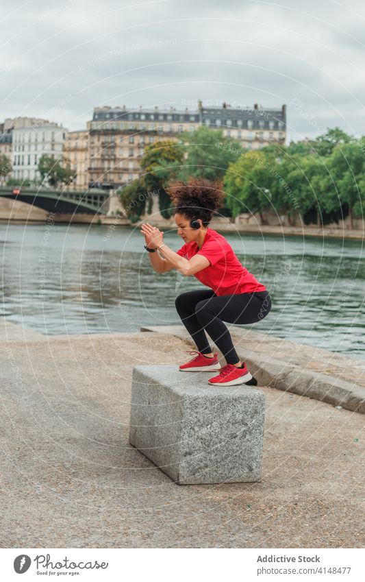 Ethnic female athlete squatting on stone during training near pond sportswoman headset warm up exercise balance river city sportswear workout activewear