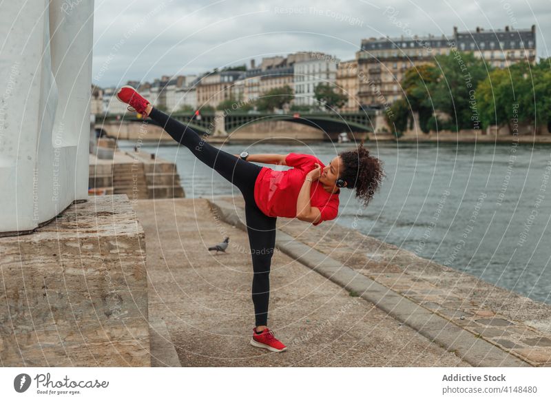 Ethnic athlete doing karate kick on pavement near city river sportswoman training workout fit flexible balance smart watch endurance determine warm up energy