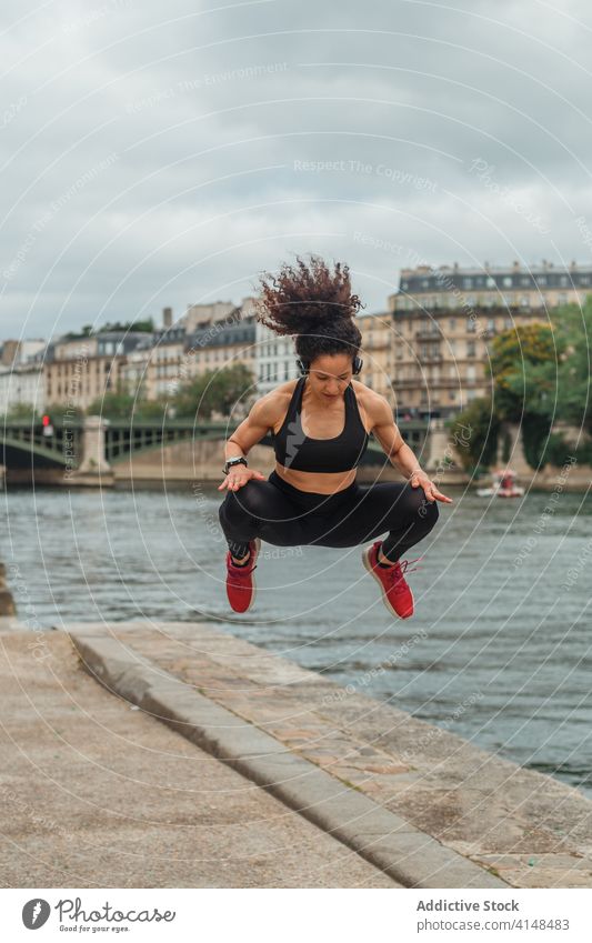 Muscular ethnic sportswoman jumping during workout near city river warm up training energy muscular fit smart watch endurance motion determine athlete headset