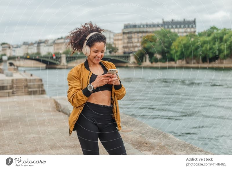 Sportswoman in headset using social media on smartphone after workout sportswoman headphones chatting internet break embankment river gadget device online