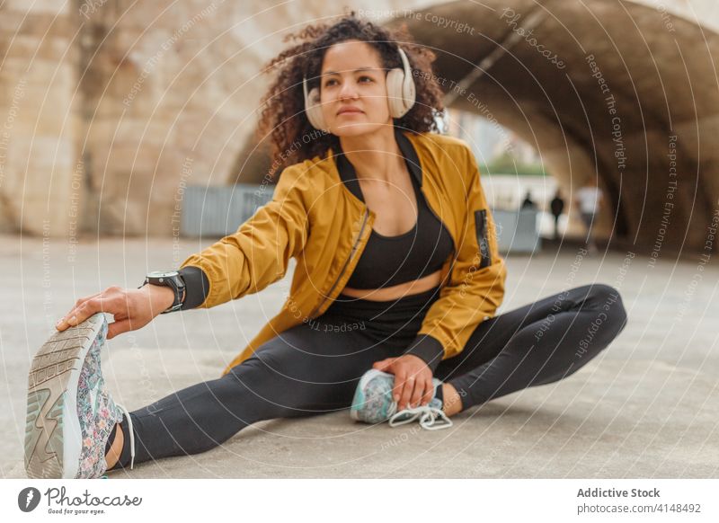 Ethnic sportswoman in headphones stretching leg on pavement in city headset exercise workout sportswear urban using device gadget smart watch asphalt road