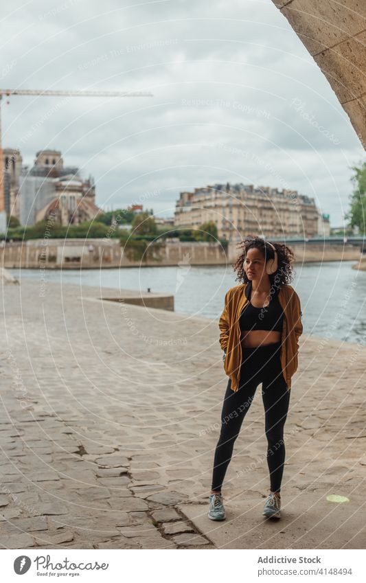Thoughtful ethnic sportswoman in headset standing near river after workout athlete headphones listen music wireless sportswear thoughtful embankment city using