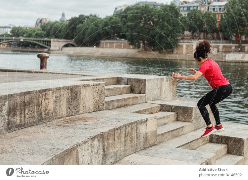 Anonymous ethnic athlete jumping on stone stairs near city pond sportswoman workout warm up training energy sportswear river cardio fit endurance motion