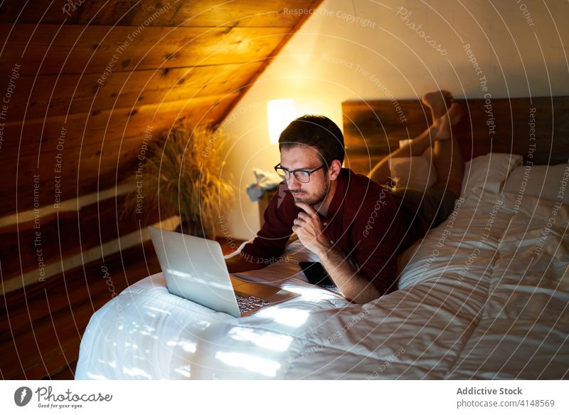Concentrated male freelancer typing on laptop at home remote man work project entrepreneur using bed lying thoughtful browsing netbook internet device gadget