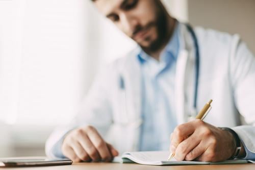 Friendly young male doctor of oriental appearance sits at a desk in his office and takes notes in a notebook, close-up medicine person health clinic stethoscope