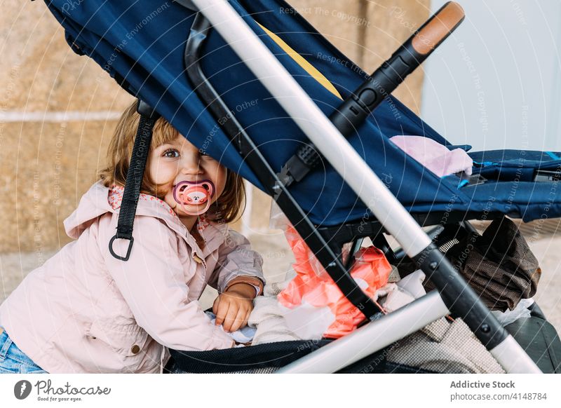 Content little kid near baby carriage baby stroller girl smile adorable street stand child cute charming joy innocent style city rest lean tender carefree relax