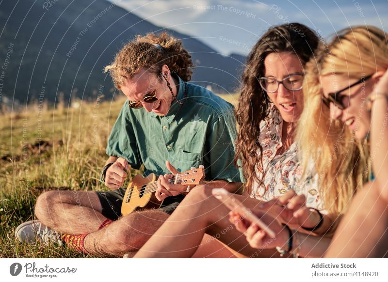 Content friends relaxing together in mountains play ukulele smartphone hipster entertain browsing friendship weekend summer grass gadget device internet joy