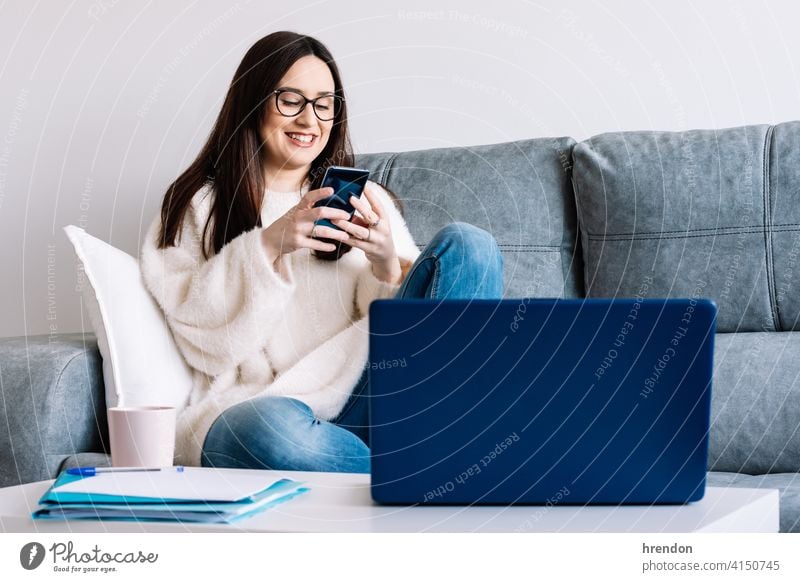 woman talking with mobile phone while working with laptop sitting on a couch at home indoor using office conversation job notebook young happy typing study