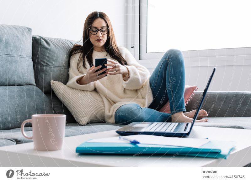 woman talking with mobile phone while working with laptop sitting on a couch at home indoor using office conversation job notebook young happy typing study