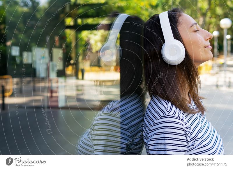 Calm woman listening to music in city headphones enjoy song carefree dreamy satisfied female glass wall building mirror reflection wireless relax happy gadget