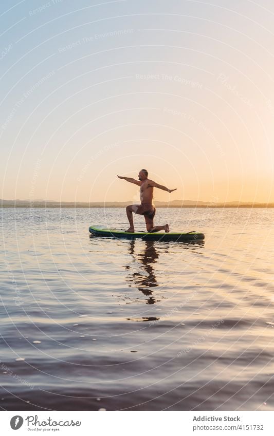 Man on paddle board standing in yoga pose on lake man practice sunset asana crescent lunge harmony anjaneyasana focus water balance float concentrate calm