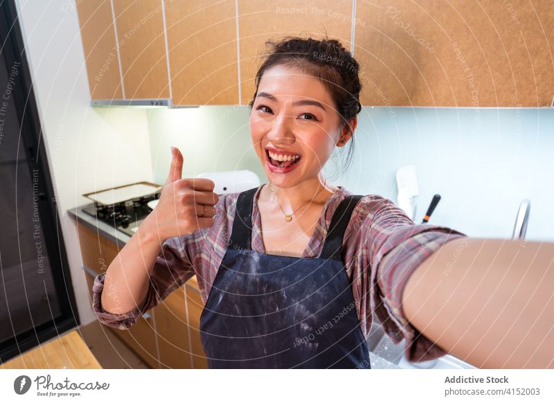 Laughing ethnic baker showing thumb up taking selfie woman home happy satisfied kitchen cheerful excited social media asian apron positive success approve laugh