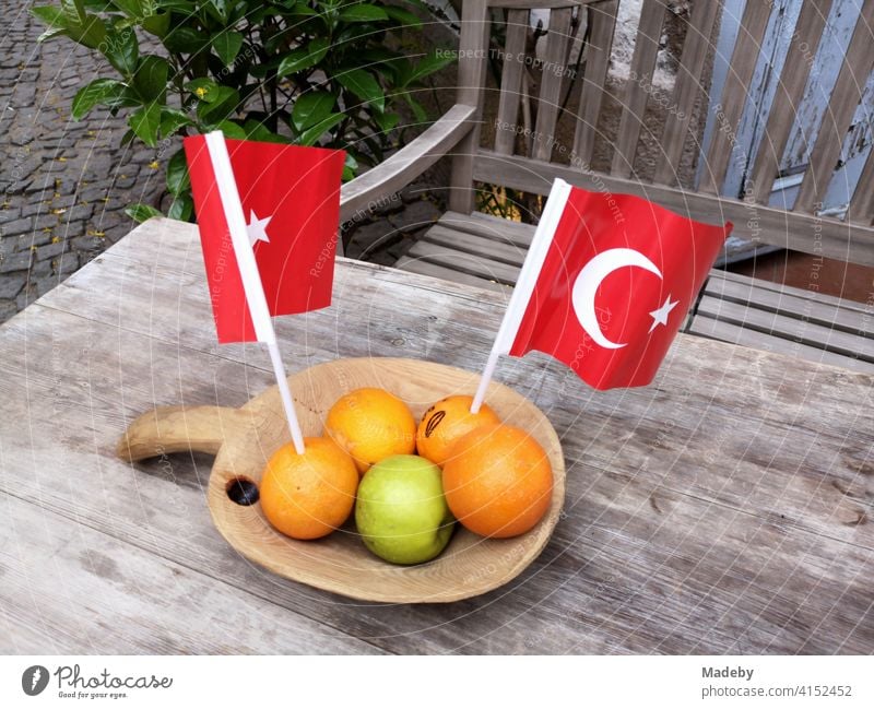 Oranges with a green apple and red Turkish pennants in a wooden bowl on an old wooden table with a wooden bench in the alleys of the old town of Alacati near Izmir in Turkey
