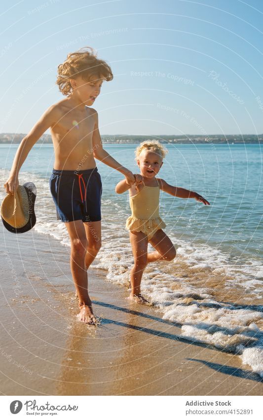 Vertical photo of two children in swimsuits holding hands playing on the seashore primary sister trust affectionate sharing feeling moment brother jump run