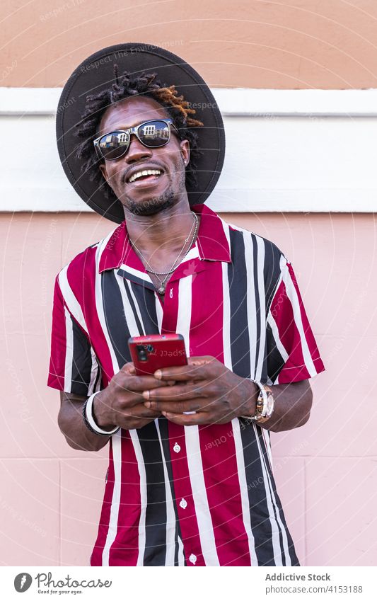 Cheerful stylish man with smartphone standing on street style fashion using trendy happy cheerful colorful sunglasses stripe hat mobile device gadget message