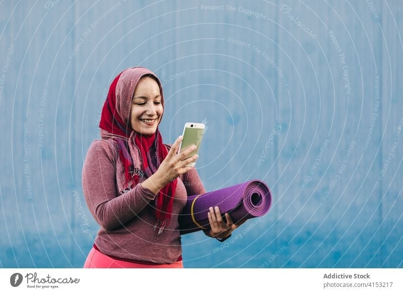 Smiling ethnic sportswoman using smartphone in city browsing hijab muslim smile mat female arab training mobile street sportswear fitness internet lady content