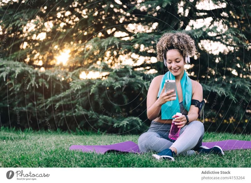 Sportive woman using smartphone after training in park sportive rest cheerful fit young browsing female headphones workout summer gadget fitness device