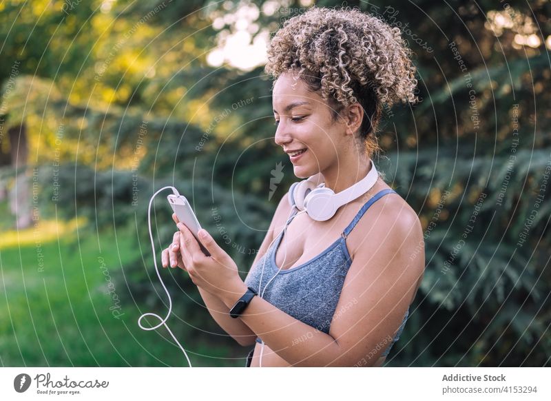Cheerful ethnic sportswoman using smartphone in park sportive rest cheerful fit young browsing chat female headphones workout summer gadget active fitness happy