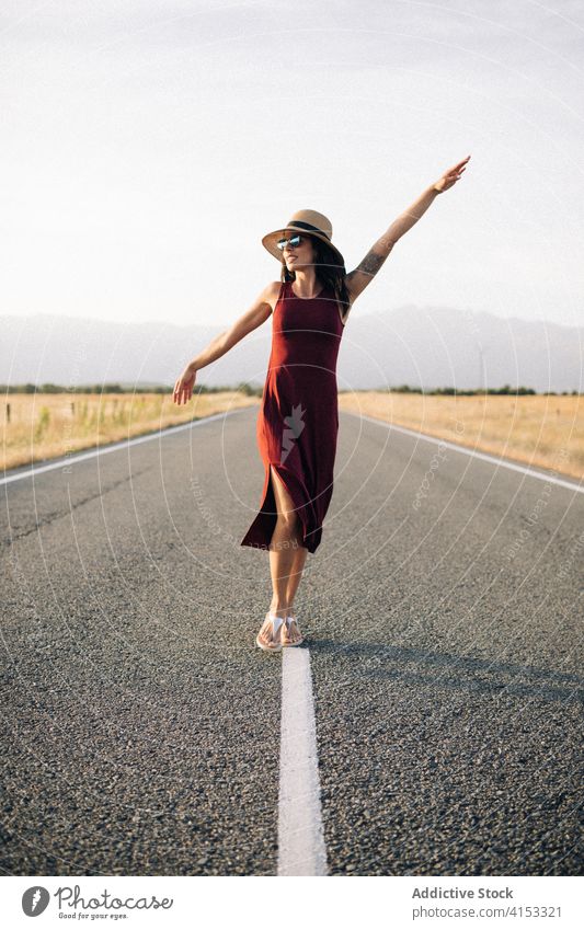 Content woman walking along road in summer carefree traveler enjoy vacation freedom smile female empty roadway dress sunhat trip happy holiday journey young