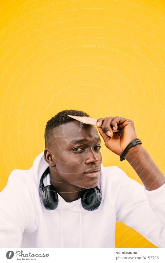 Young african American black man combing his afro hair while seated on a yellow wall background, wearing a white sweatshirt and headphones. Looking to the camera with serious expression. Copy space