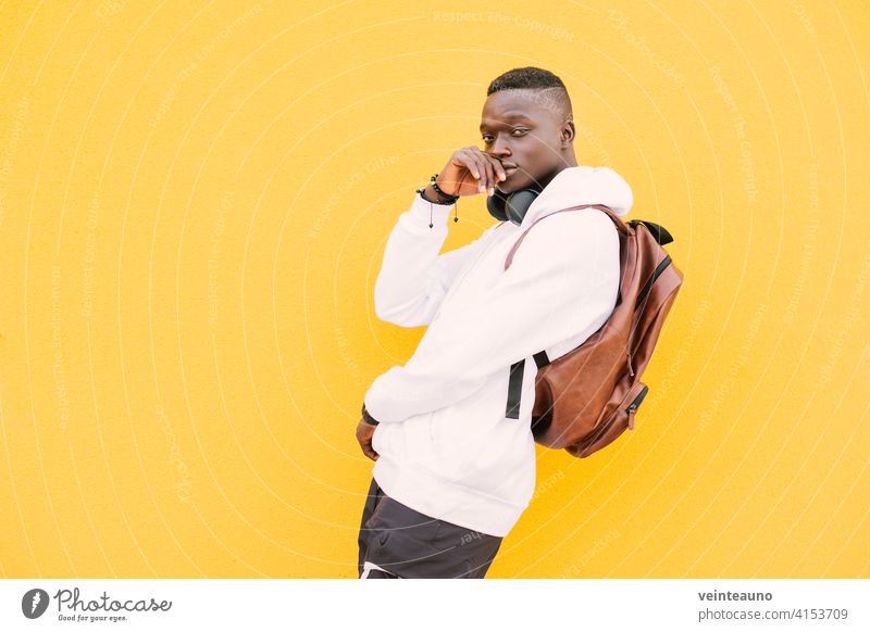 Young african American black man against a yellow wall wearing a white sweatshirt and a backpack listening music on wireless headphones while looking at camera with attitude