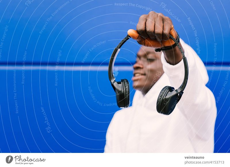 Young black man holding wireless headphones while wearing a white sweatshirt, against a blue wall looking away with confidence. Selective focus young african