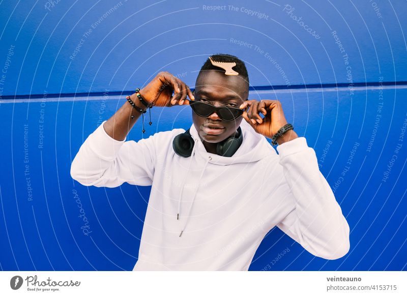 Young afro American black man wearing a white sweatshirt, sunglasses, comb and headphones on a blue wall looking over the glasses with confidence young african