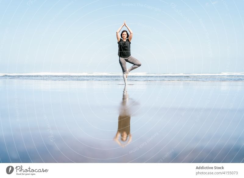 Young woman in balancing yoga asana standing on beach sea practice pose tree vrksasana balance calm seashore harmony eyes closed wellness lifestyle female