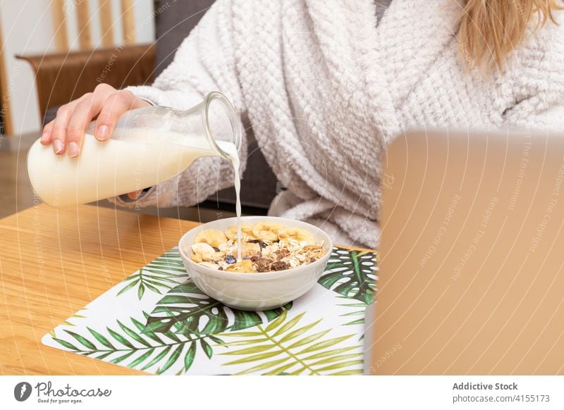 Anonymous woman having breakfast at home bowl cereal milk pour morning nutrition healthy food female delicious table laptop tasty bathrobe positive meal fresh