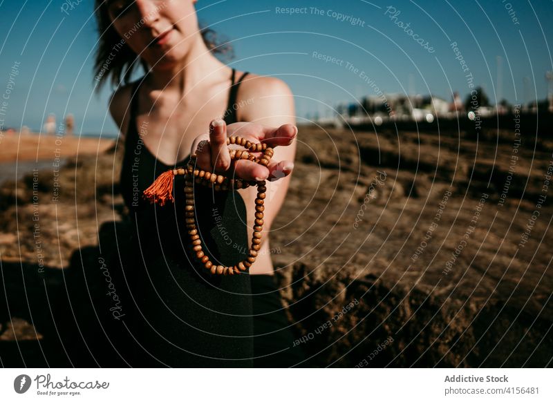 Crop woman with rosary doing yoga on beach asana practice bead prayer pose calm flexible female rocky shore healthy tranquil slim serene zen relax position