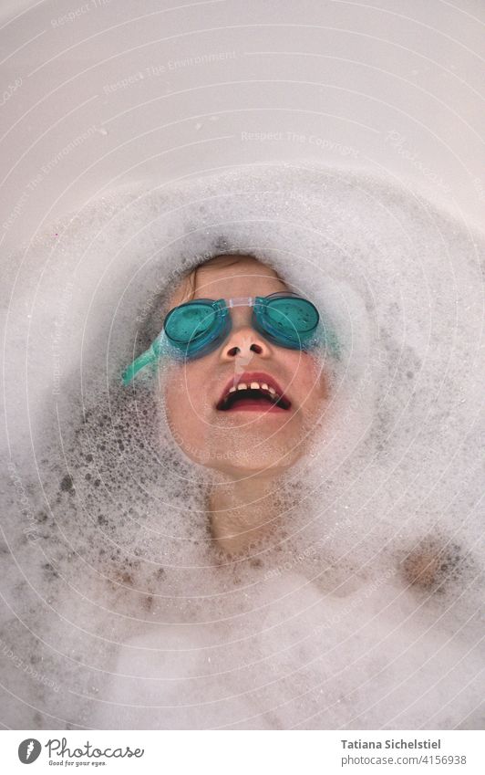 child lies in the bathtub with turquoise swimming goggles, only the face is visible bathe Bathtub Child Diving goggles Dive Foam Turquoise Water Face Laughter