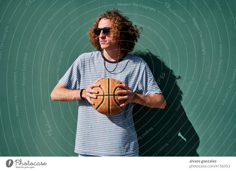 a young man with a basketball ball and sunglasses with a green wall behind looking at the sun che person sport athletic male game player competitive background